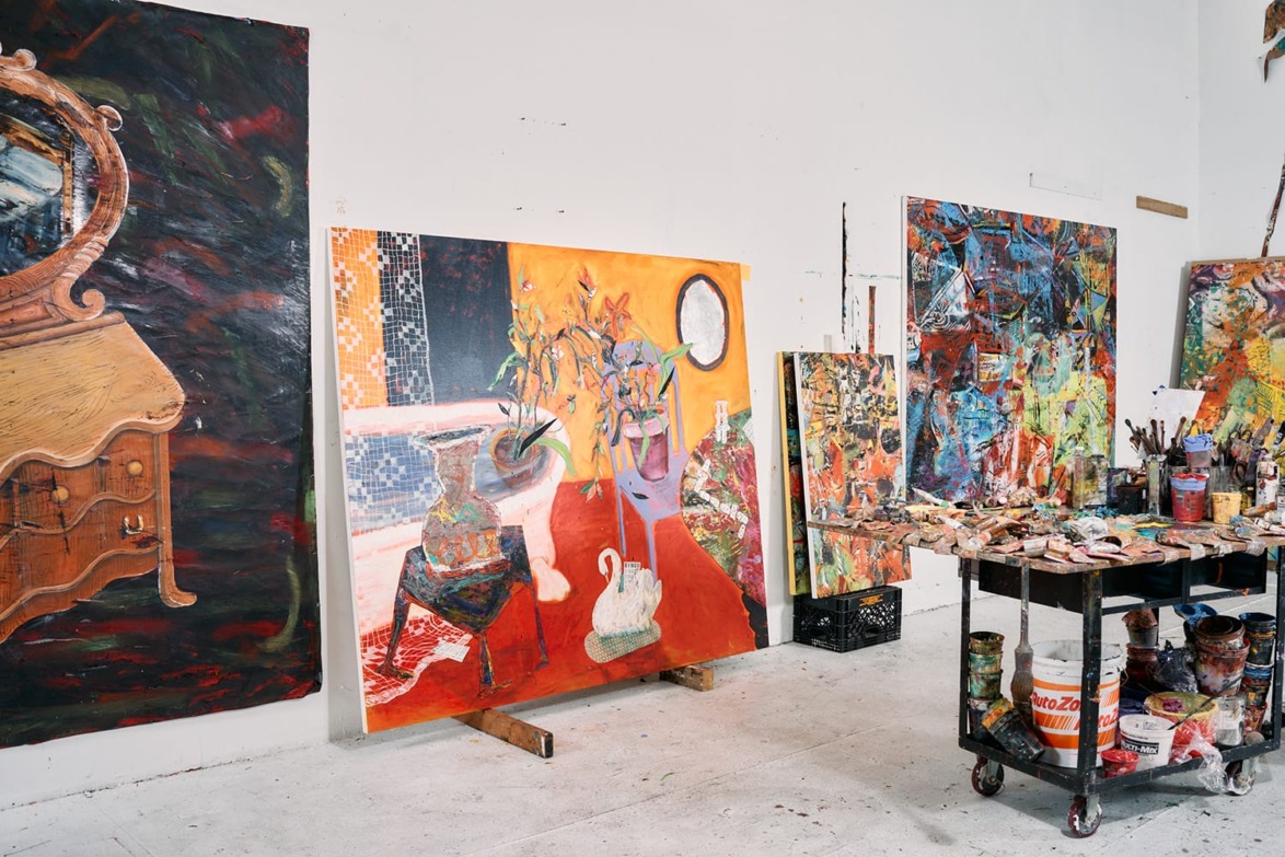 Art studio with colorful paintings, a wooden art cart, and various paints and brushes on a light gray floor.