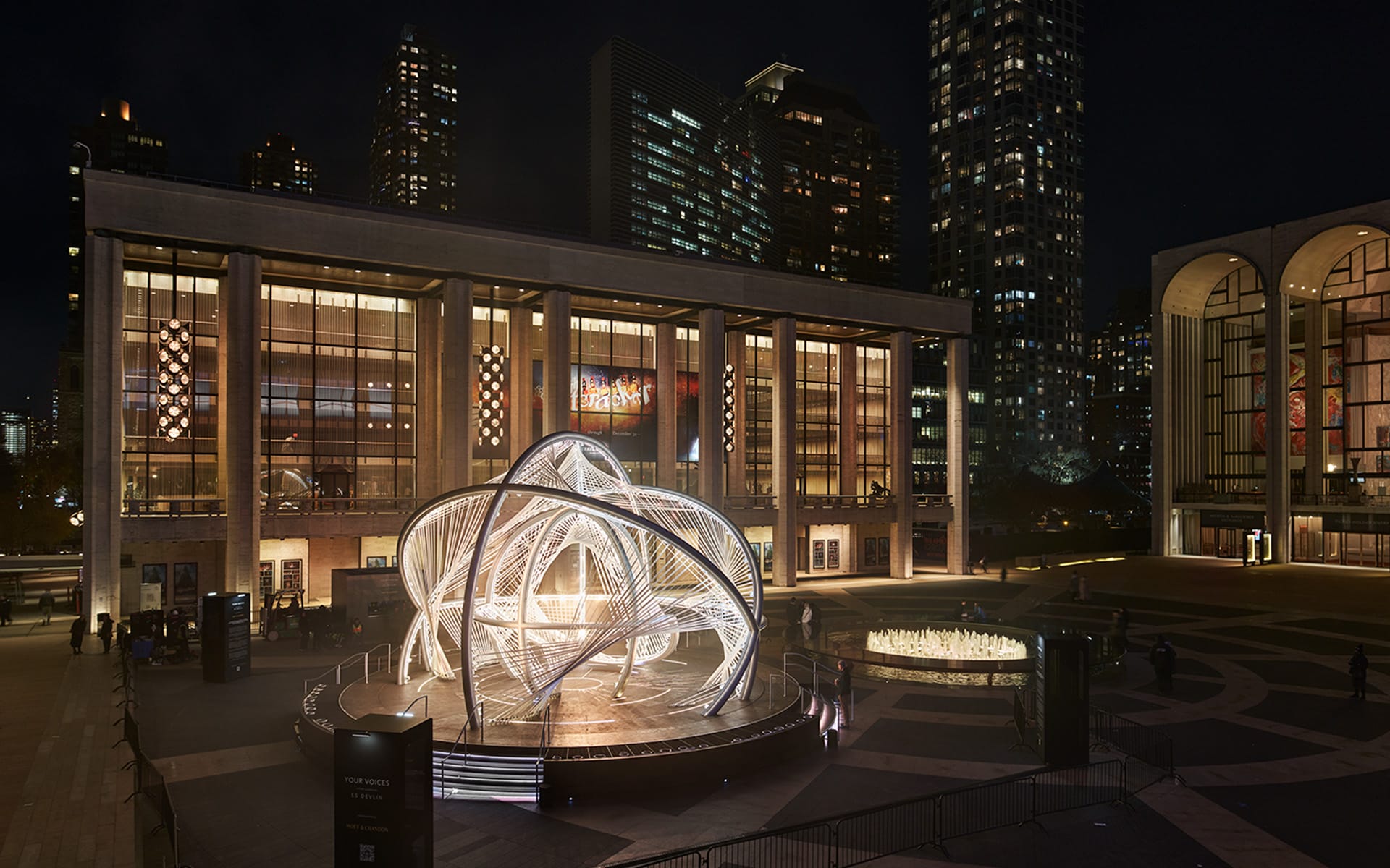 Lincoln Center at night with a modern illuminated sculpture in the foreground and surrounding city lights.