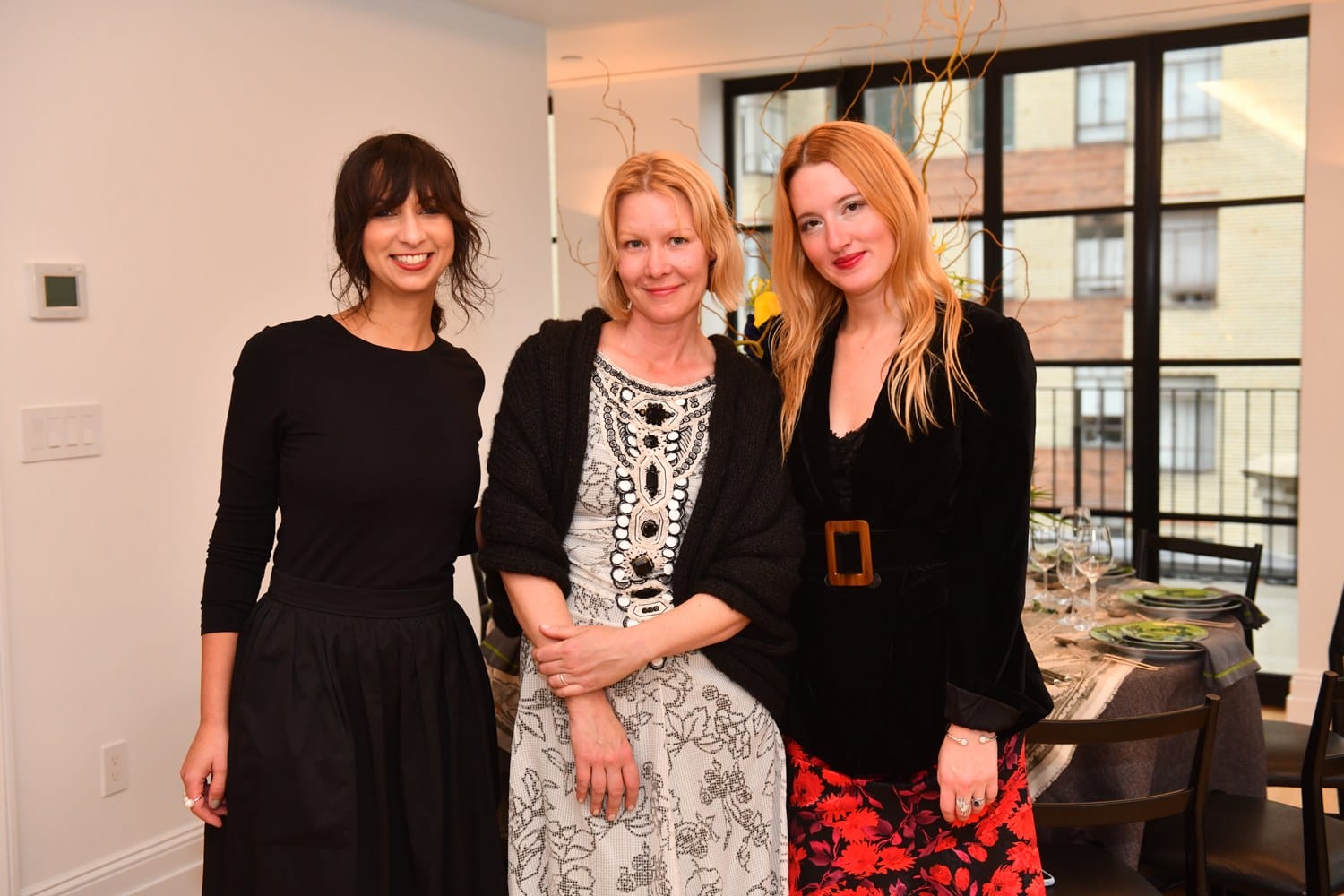 Three women are standing together indoors, smiling, with a table set for dinner in the background.