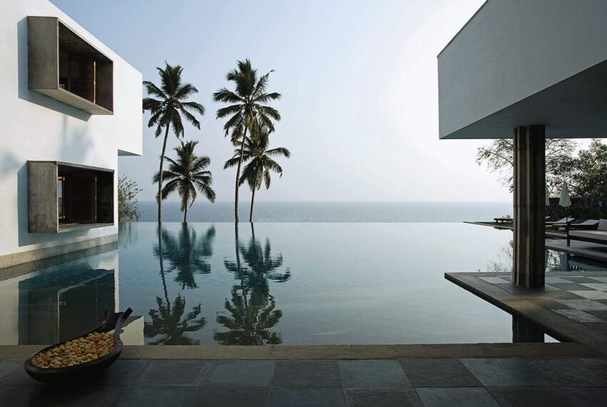 Infinity pool with ocean view, surrounded by modern architecture and palm trees, under a clear blue sky.
