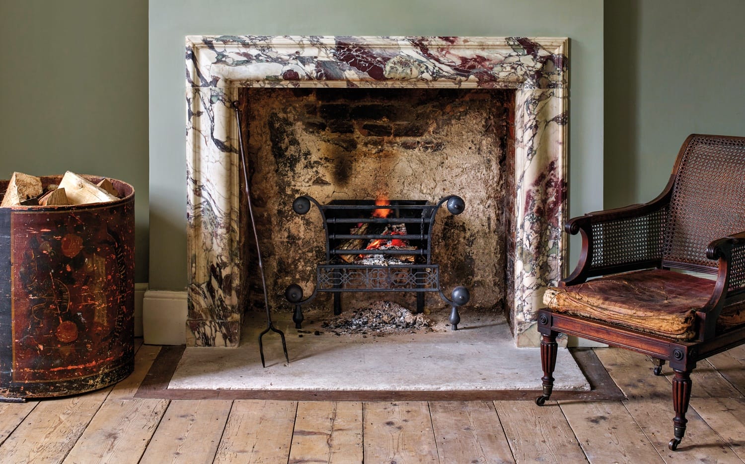 Marble fireplace with lit fire, vintage chair, and wood basket on wooden floor in a cozy room.