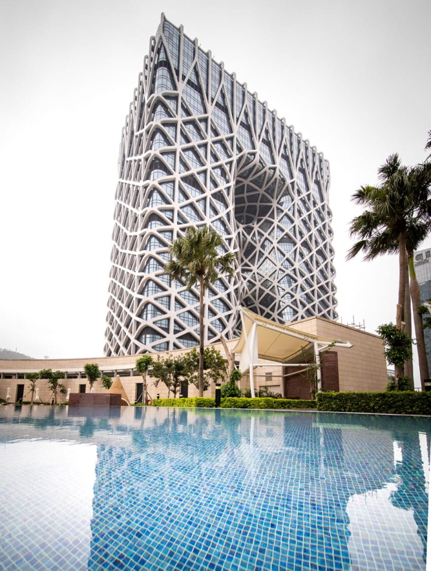 Modern high-rise building with unique geometric design reflected in a swimming pool, surrounded by palm trees and greenery.