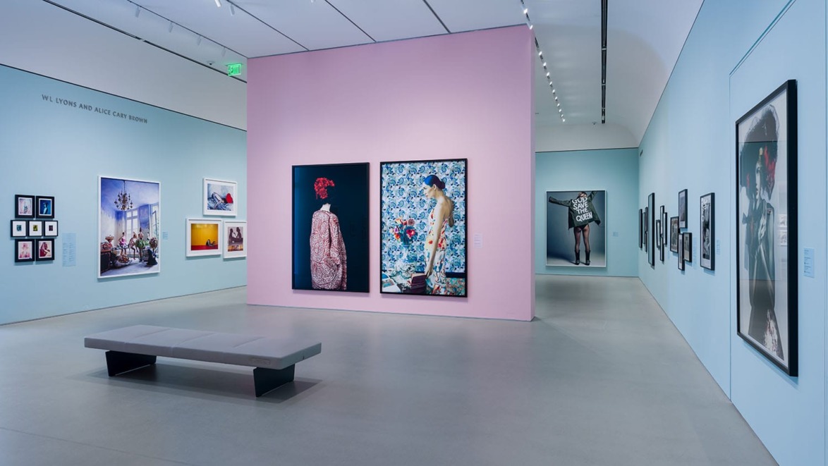 Modern art gallery interior with vibrant photographs and paintings displayed on pink and blue walls, and a bench in the center.