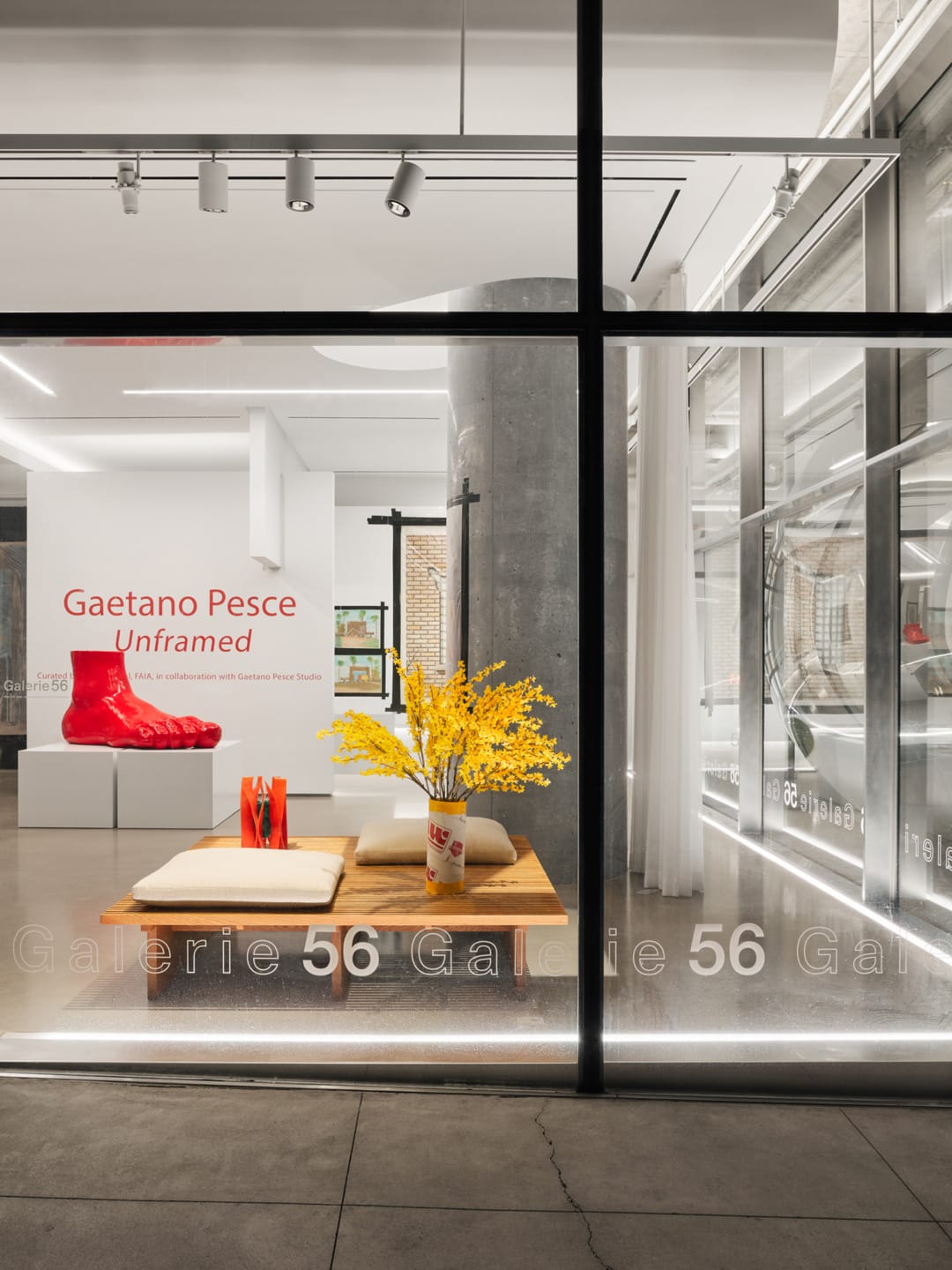 Gallery window display with modern art: large red shoe sculpture, yellow flowers in a vase, and minimalist interior design.