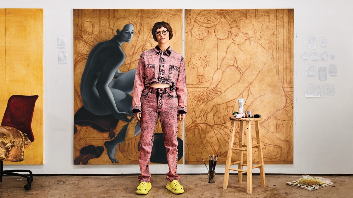Artist in unique outfit standing in front of large canvas paintings, with art supplies and sketches visible in studio setting.