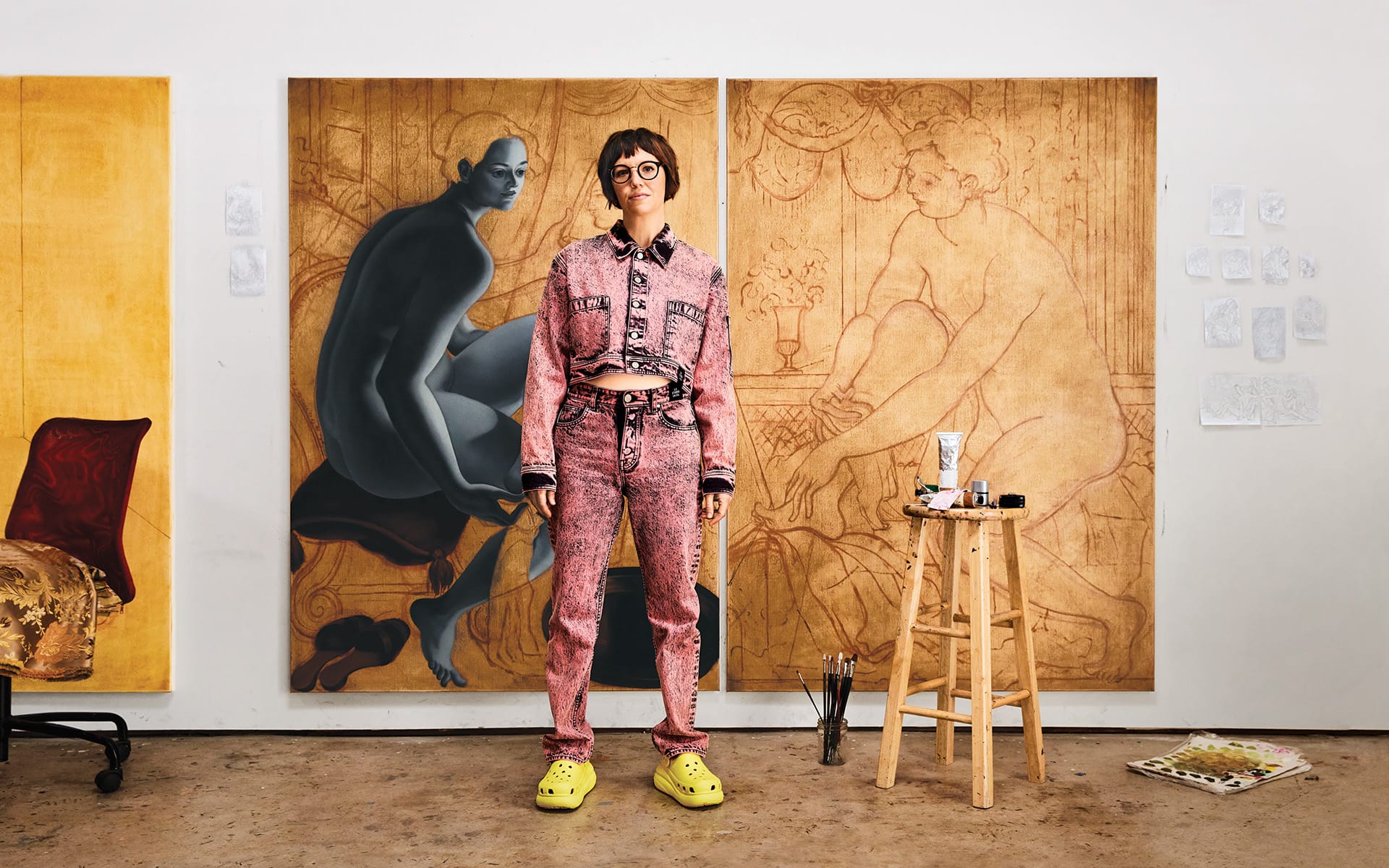Artist in unique outfit standing in front of large canvas paintings, with art supplies and sketches visible in studio setting.