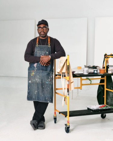 Artist in a studio wearing an apron, standing next to a cart with paint supplies, neutral background.