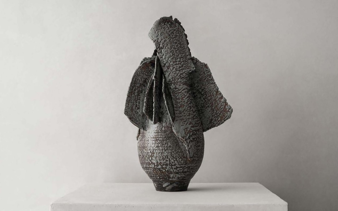 Textured, dark ceramic sculpture with layered, wing-like structures on a pedestal against a plain background.