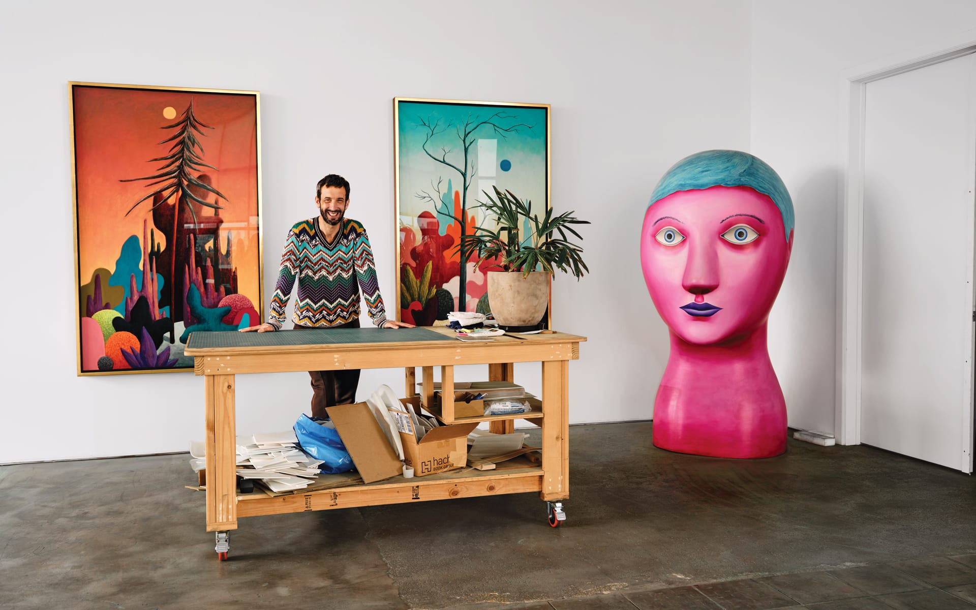 Artist in colorful sweater standing by a worktable in a studio with vibrant paintings and a large pink sculpture head.