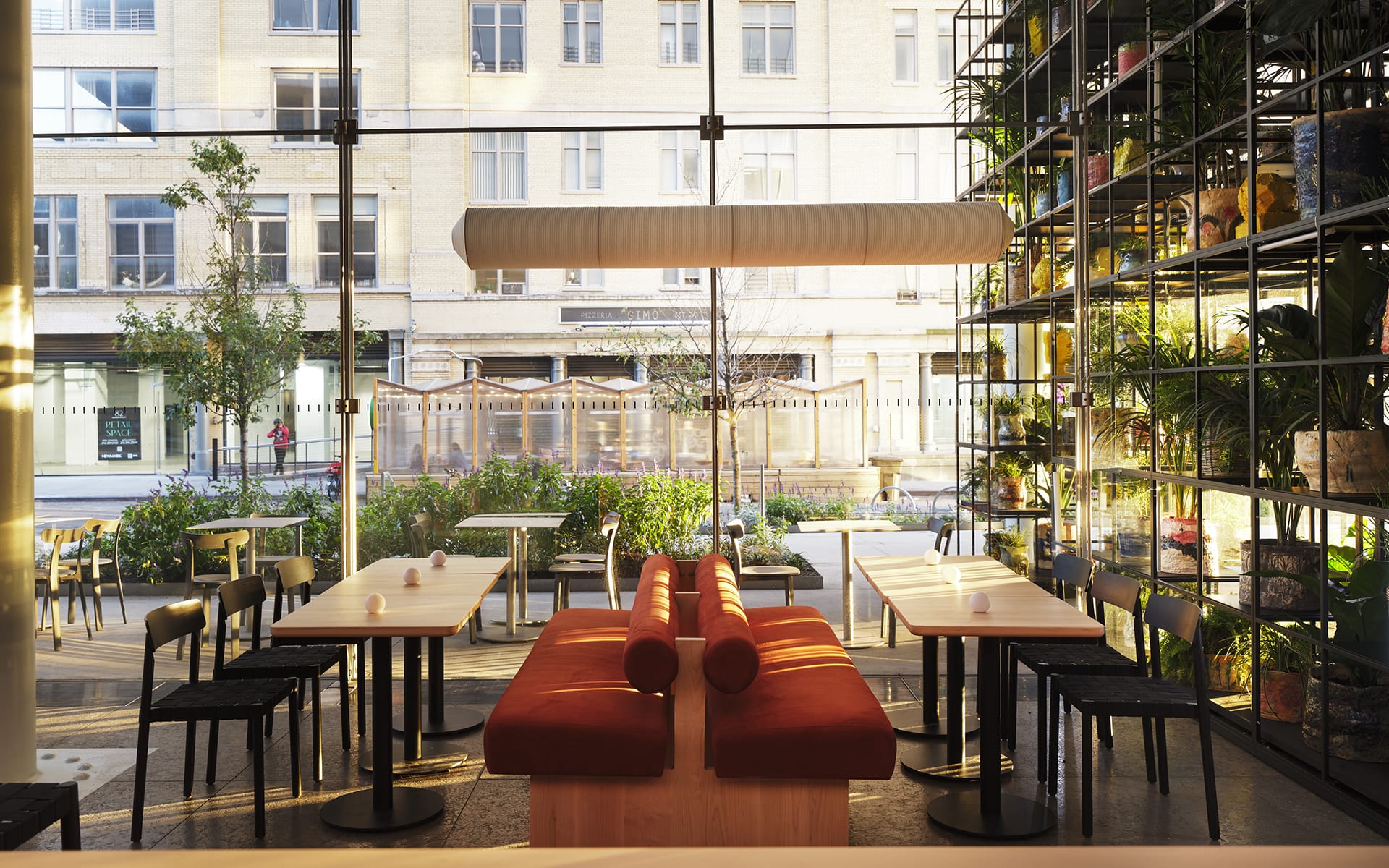 Modern cafe interior with sunlight streaming through large windows, featuring plants, wooden tables, and red cushioned seating.