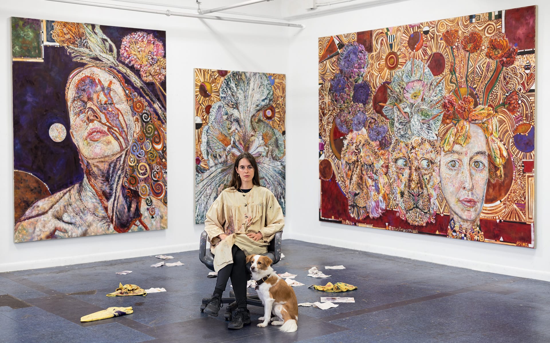 Artist sitting in a gallery surrounded by colorful, abstract paintings with a dog resting nearby on the floor.