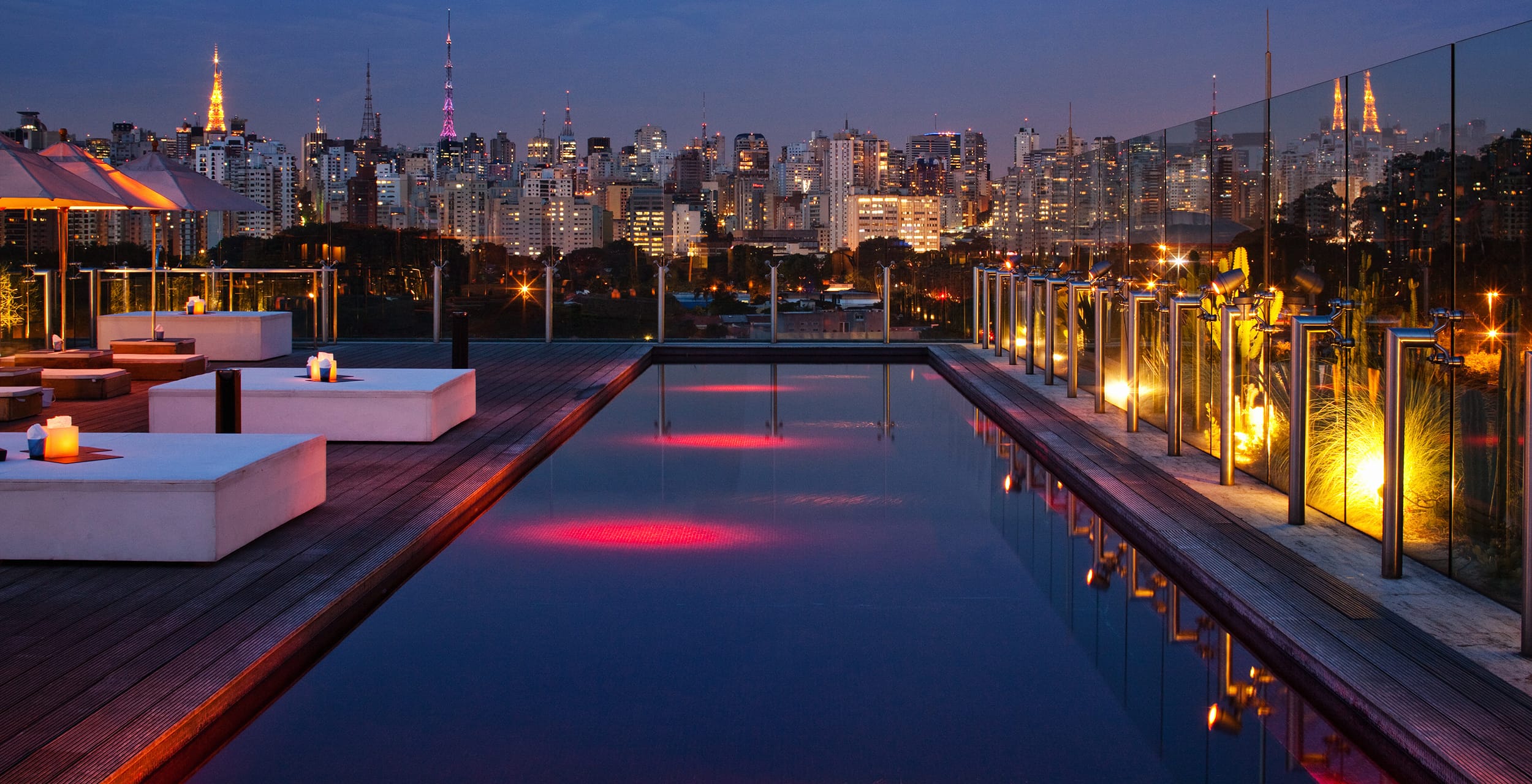 Rooftop pool with glowing lights, surrounded by modern seating, overlooking a city skyline at dusk.
