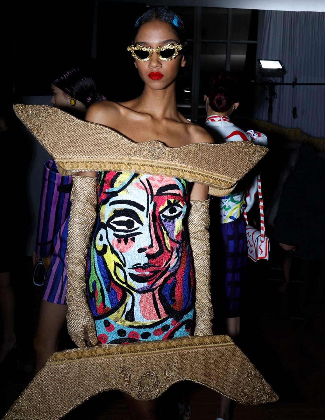 Model in avant-garde dress with colorful abstract face design and ornate gold accessories on runway.