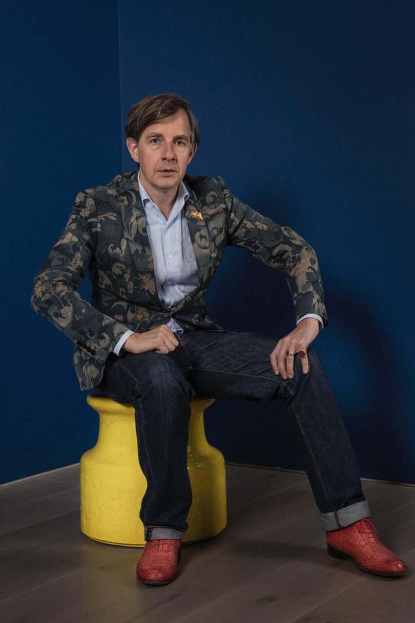 Person sitting on yellow stool against blue wall, wearing a floral jacket, blue shirt, jeans, and red shoes.