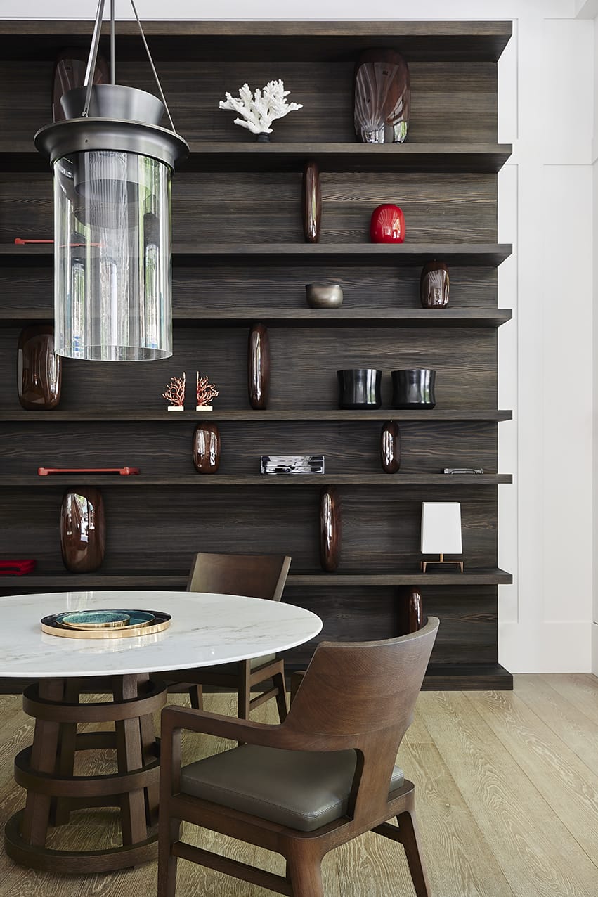 Modern dining area with a round table, brown chairs, pendant light, and decorative shelves on a wooden accent wall.