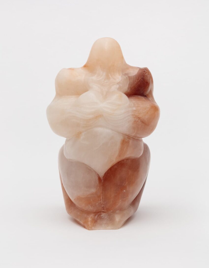 Abstract stone sculpture of a nude figure with folded arms, showcasing smooth curves and warm earthy tones on a neutral background.