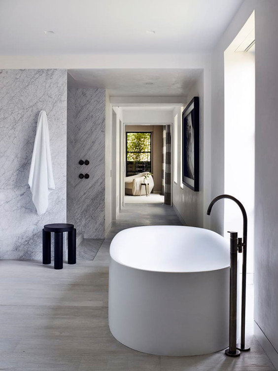Modern bathroom with freestanding oval tub, marble wall, black stool, and large window with natural light.