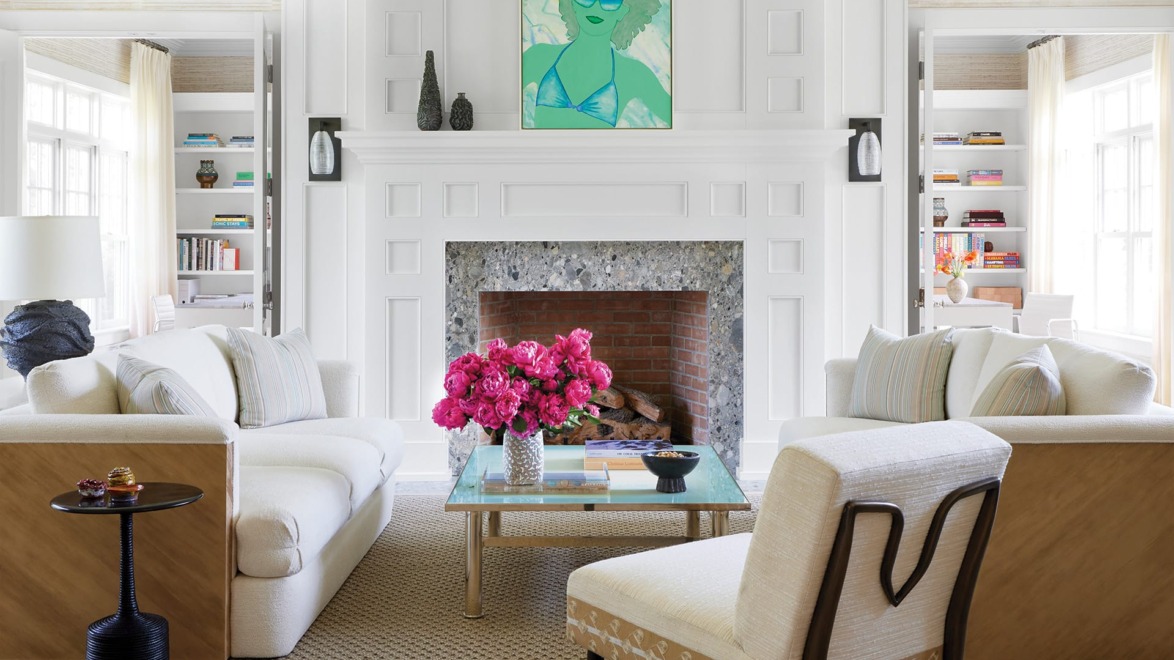 Bright living room with white sofas, a brick fireplace, vibrant artwork, pink flowers on a glass coffee table, and bookshelves.