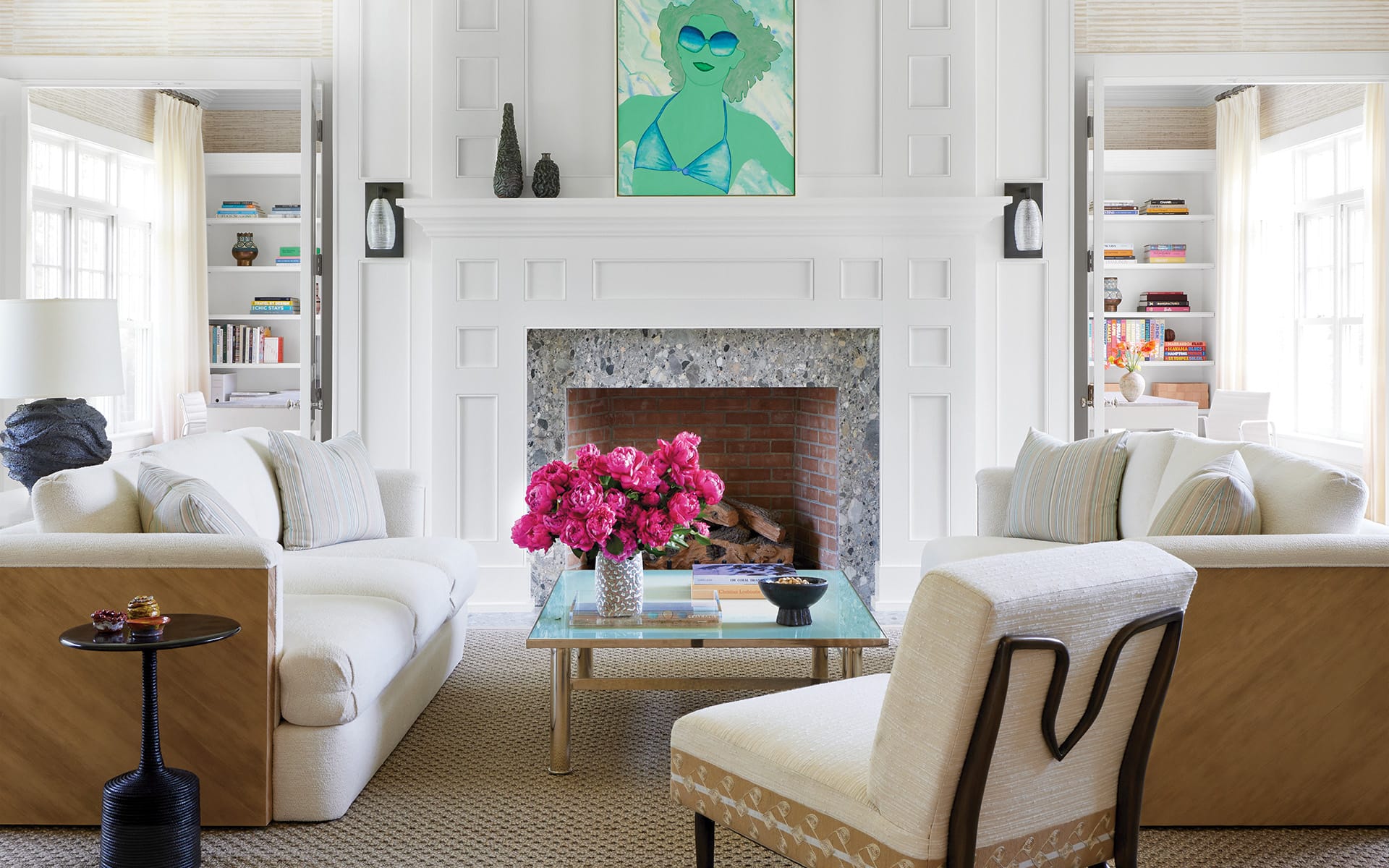 Bright living room with white sofas, a brick fireplace, vibrant artwork, pink flowers on a glass coffee table, and bookshelves.
