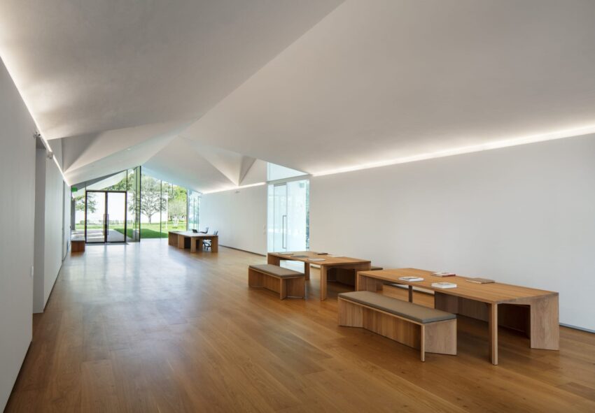 Modern, minimalistic interior with wood flooring featuring wooden tables and benches, overlooking a garden through glass walls.