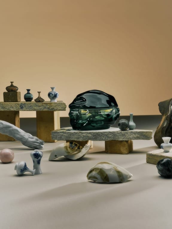 Artistic arrangement of various miniature sculptures and a dark green glass object on textured stone platforms against a beige backdrop.