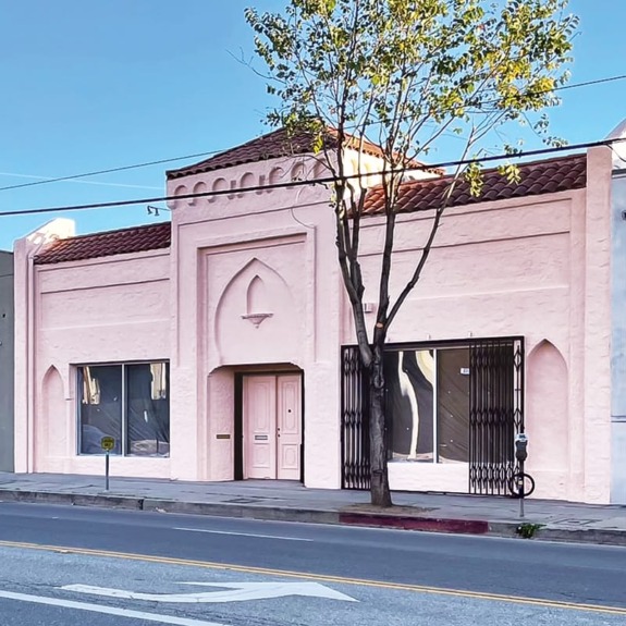 Pink stucco building with arched doorway, large windows, and a tree in front, set on an empty street under a clear sky.