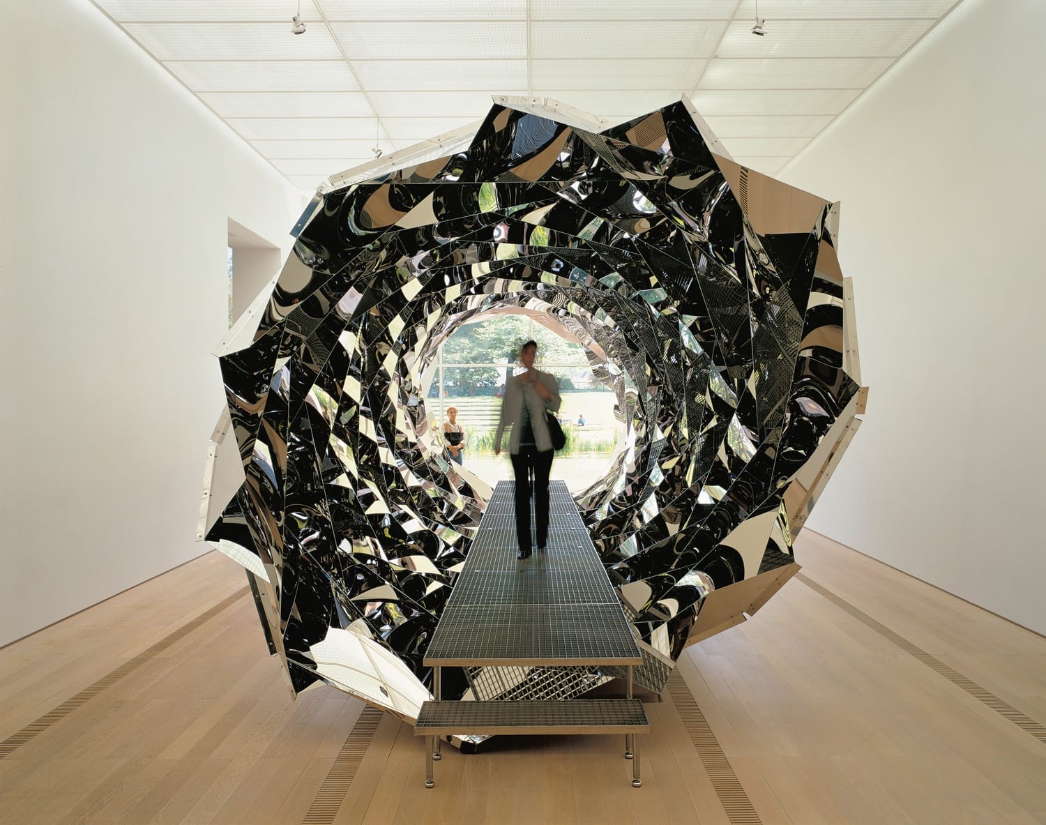 Person walking through a reflective, geometric tunnel in a modern art gallery setting with wooden floors.