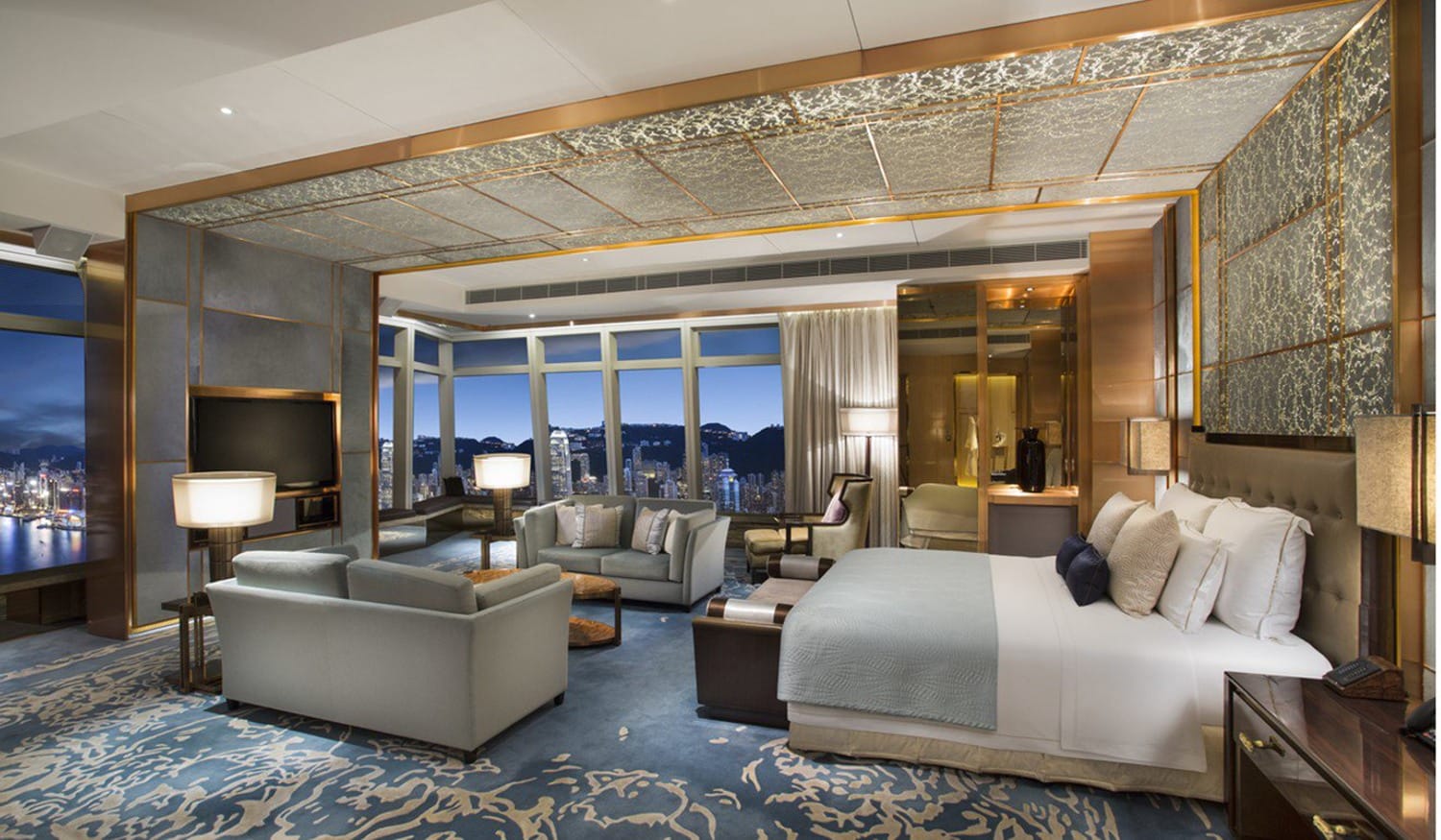 Luxurious hotel room with a large bed, seating area, and a city view through floor-to-ceiling windows at night.