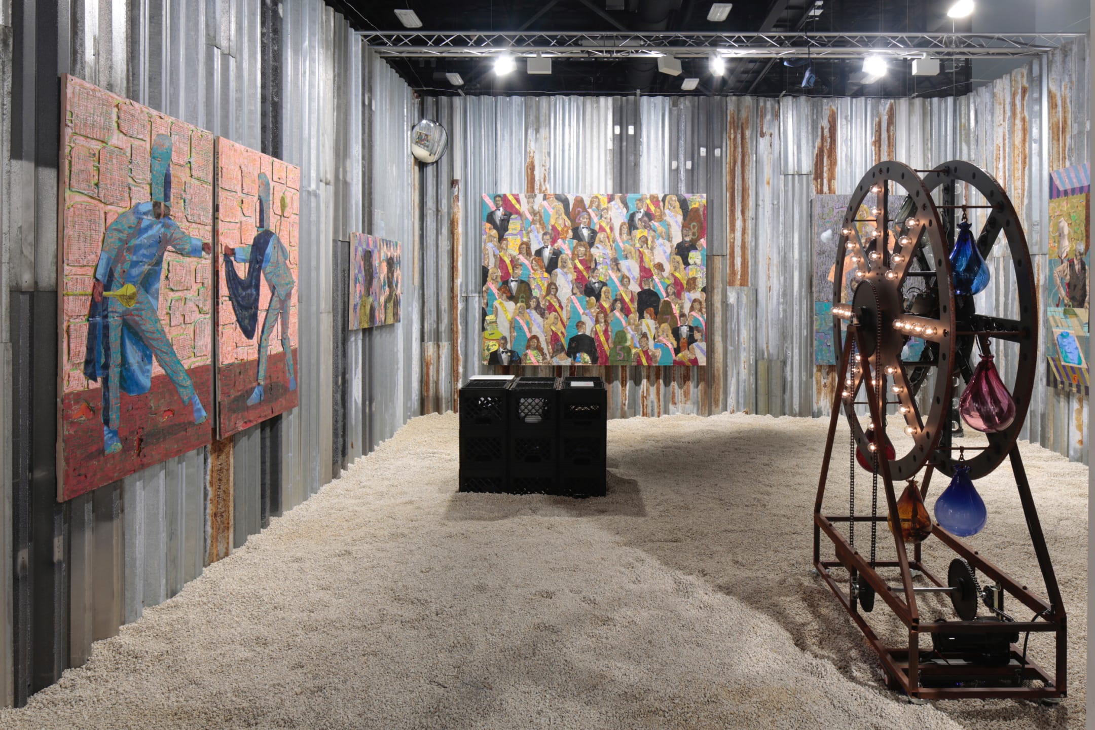 Art installation with colorful paintings on corrugated metal walls, small black crates, and a wheel with suspended glass shapes.