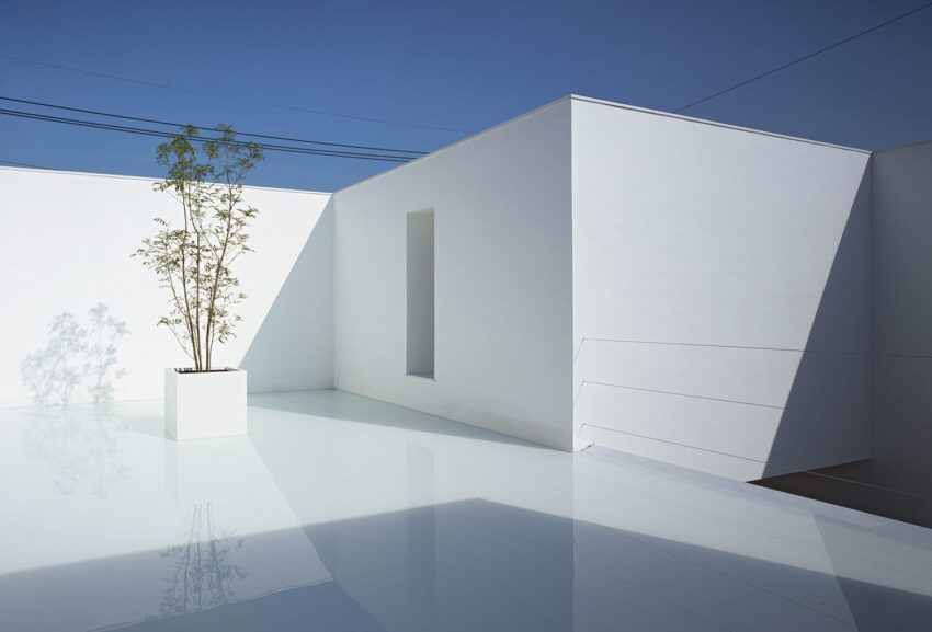 Minimalist white building with a solitary potted tree, reflecting on a polished surface under a clear blue sky.
