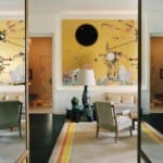 Modern living room with abstract artwork, mirrored wall, beige chairs, and yellow-accented rug, creating a stylish ambiance.