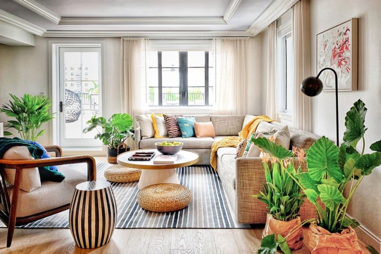 Modern living room with large windows, beige sofa, indoor plants, striped rug, and round coffee table with decorative items.