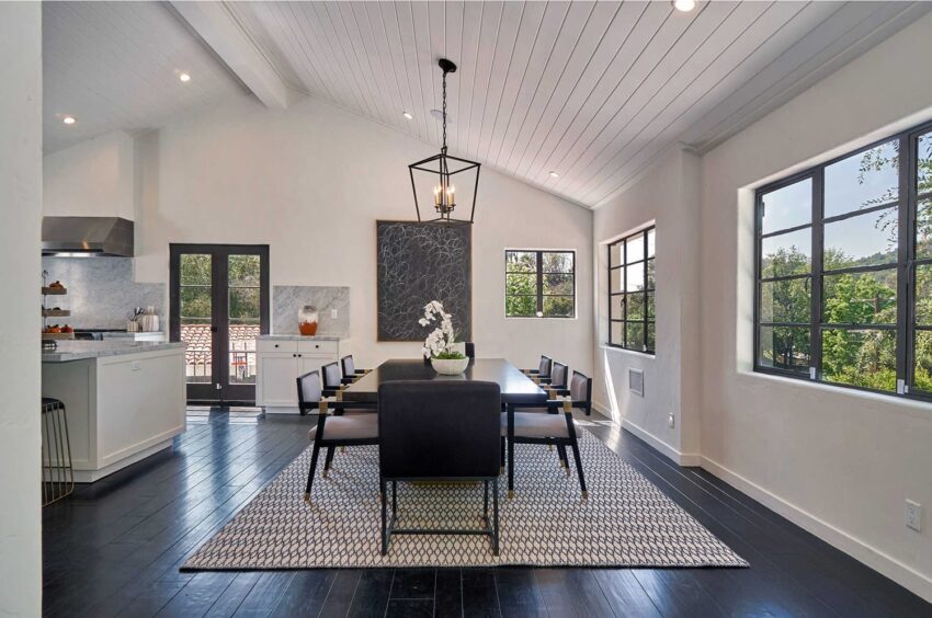 Modern dining room with dark wood flooring, large table, black chairs, white walls, and windows overlooking greenery.