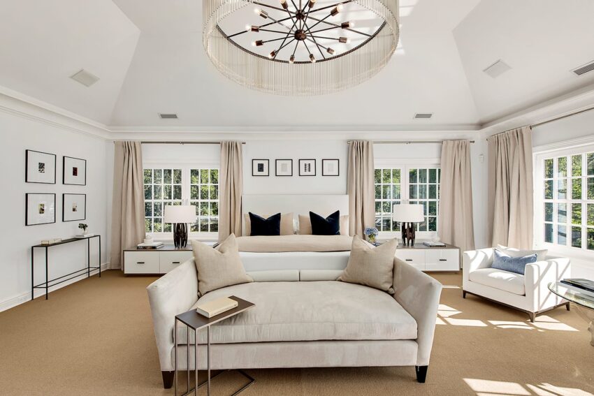 Spacious bedroom with beige sofa, modern chandelier, and large windows with natural light streaming in.