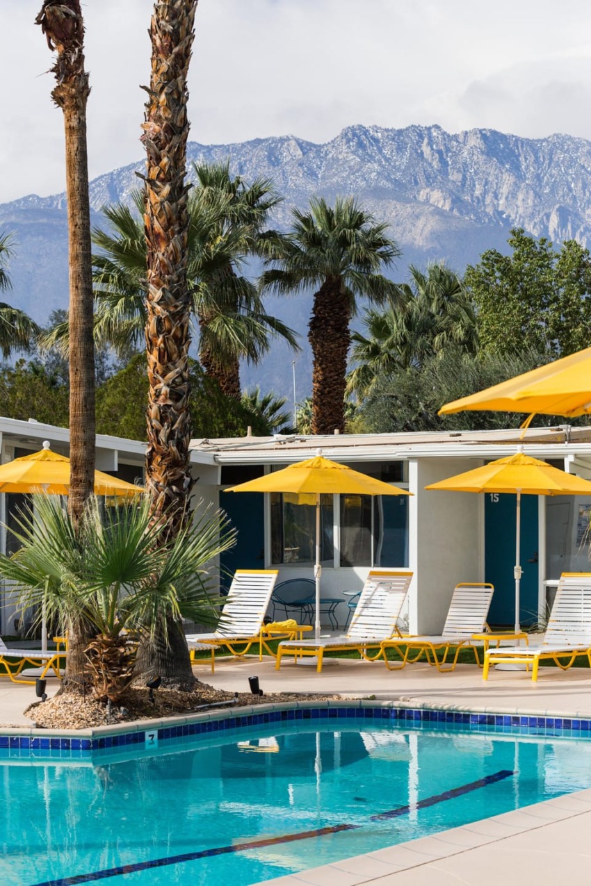 Retro resort pool area with yellow umbrellas, palm trees, and a mountain backdrop under cloudy skies.