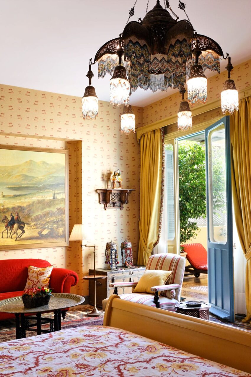 Elegant vintage bedroom with ornate chandelier, colorful furniture, and open door leading to a sunlit patio with greenery.