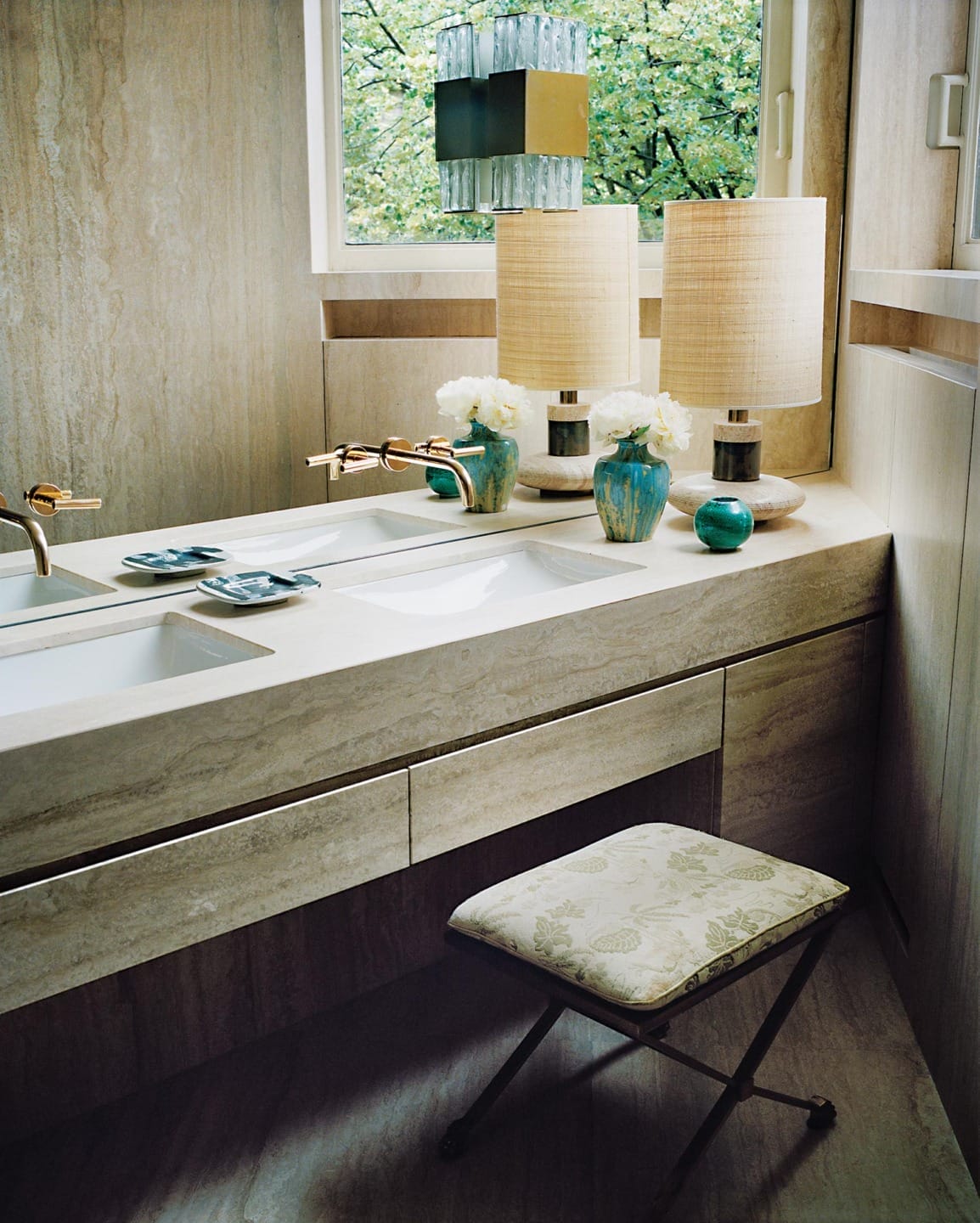 Modern bathroom with dual sinks, beige marble countertop, decorative lamps, green vases, and a patterned stool.