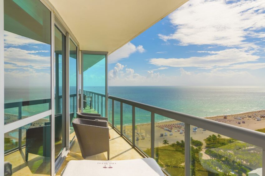 Ocean view from a high-rise balcony with glass railings, wicker chairs, and beach in the distance under a clear blue sky