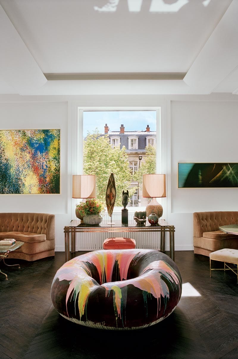 Elegant living room with large abstract chair, vibrant paintings, and a view of a city street through a large window.