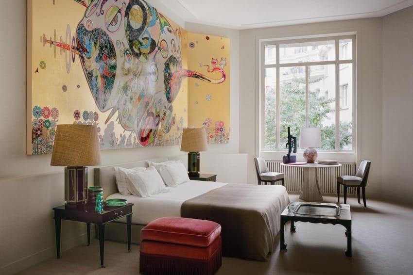 Modern bedroom with large abstract wall art, double bed, side tables with lamps, large window, chairs, and a small table.