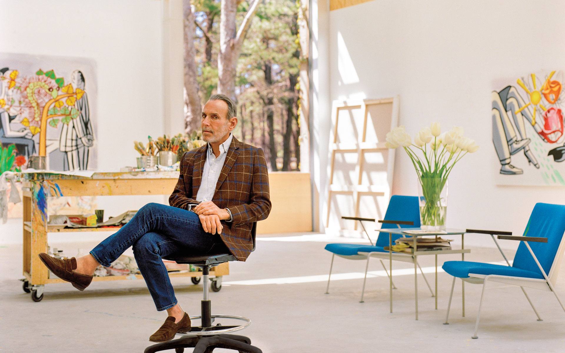 Man sitting on chair in art studio with paintings, art supplies, and plants surrounding. Bright and spacious interior.