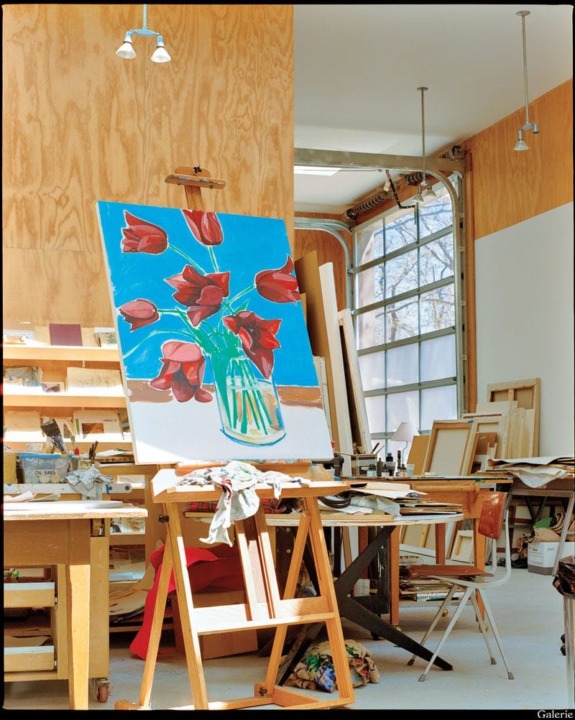 Art studio with a painting of red tulips on an easel, surrounded by shelves, framed artwork, and large windows.