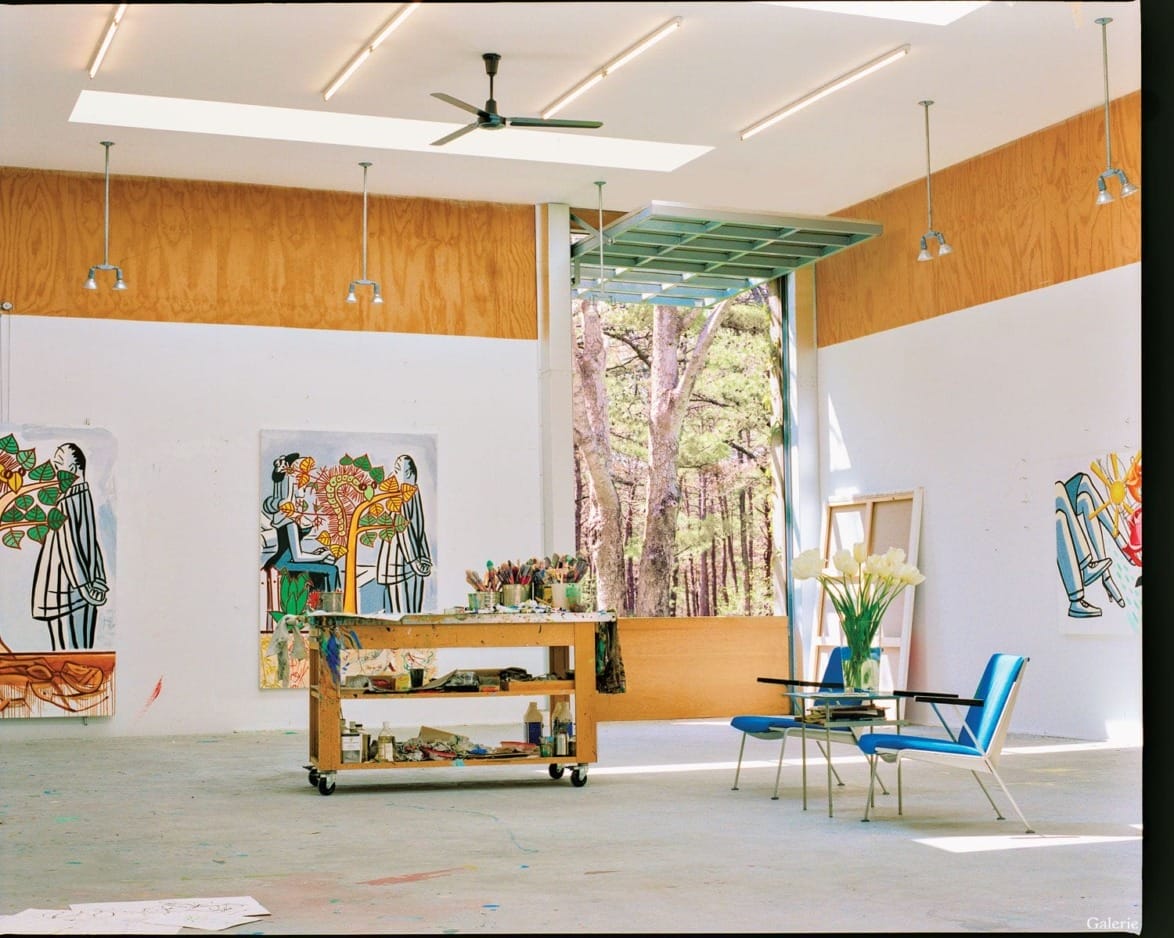 Spacious art studio with large open doors, paintings on walls, art supplies on a trolley, and chairs with a table.