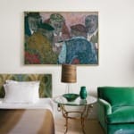 Bedroom with green armchair, round glass table, lamp, and colorful abstract painting above the bed.