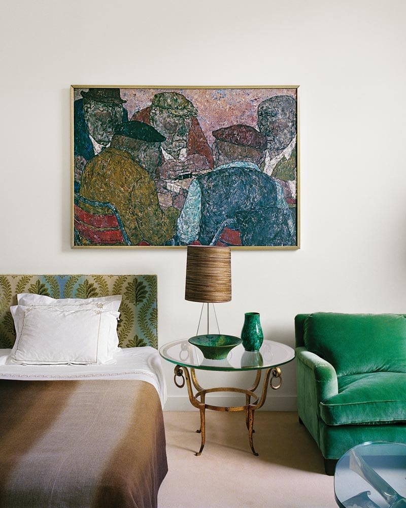 Bedroom with green armchair, round glass table, lamp, and colorful abstract painting above the bed.
