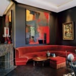 Modern living room with red velvet sofa, abstract wall art, dark walls, marble fireplace, and eclectic decor.