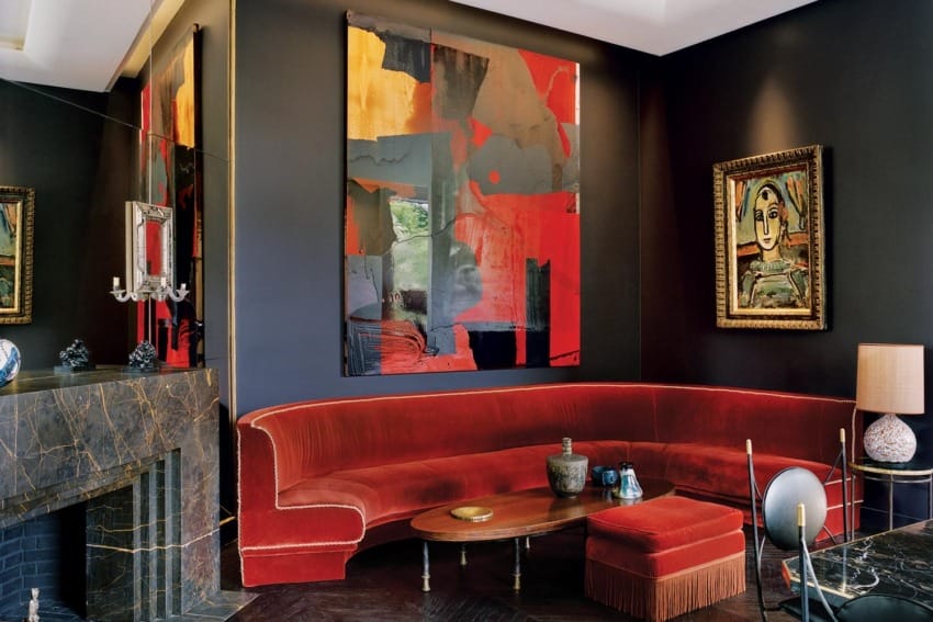 Modern living room with red velvet sofa, abstract wall art, dark walls, marble fireplace, and eclectic decor.