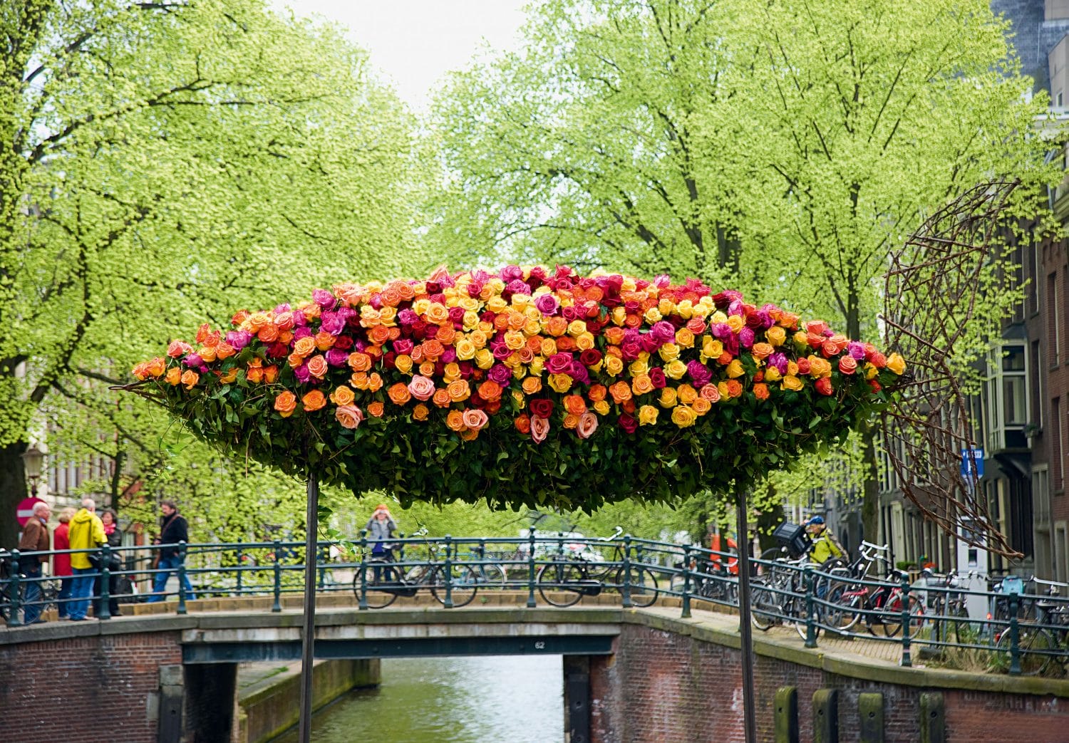 Colorful flower arrangement resembling a boat over a canal bridge surrounded by lush green trees and bicycles.