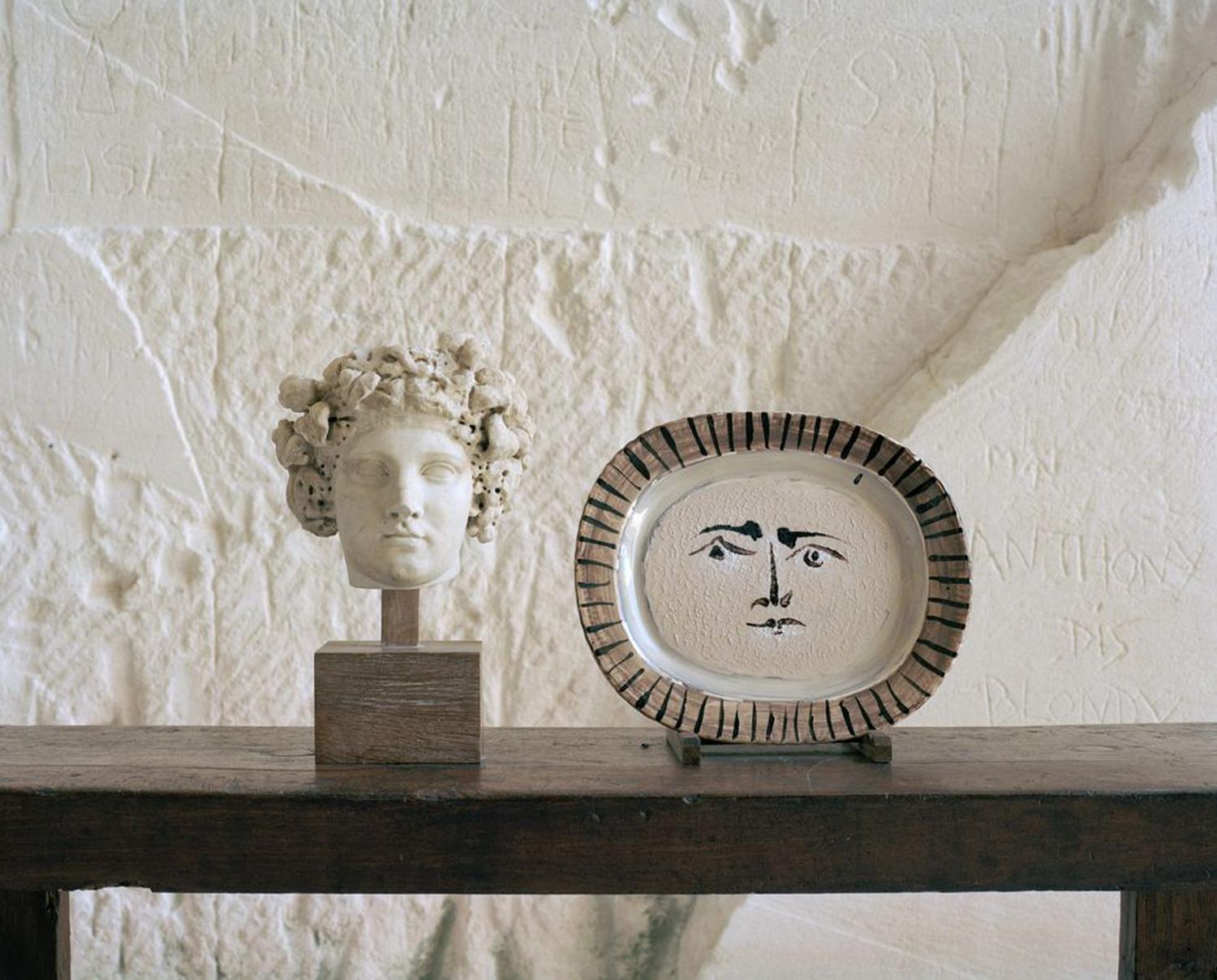 Bust of a classical head next to a decorative plate with a painted face on a wooden table against a textured wall.