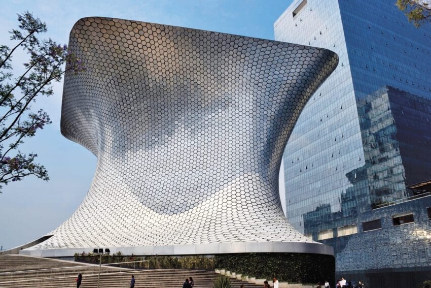 Modern architecture of a silver, hexagon-patterned museum building against a blue sky with a glass building in the background.